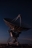 A large satellite dish antenna against the backdrop of a dark evening or night sky. The structure is illuminated by subtle lighting, highlighting its intricate metal framework and mechanical parts.