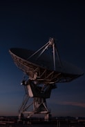 A large satellite dish antenna against the backdrop of a dark evening or night sky. The structure is illuminated by subtle lighting, highlighting its intricate metal framework and mechanical parts.
