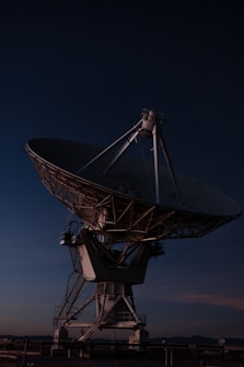 A large satellite dish antenna against the backdrop of a dark evening or night sky. The structure is illuminated by subtle lighting, highlighting its intricate metal framework and mechanical parts.