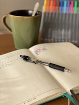 An open notebook with handwritten notes is lying on a wooden surface. A black pen rests diagonally across the notebook. To the left, a green mug with a spoon inside is positioned, and to the right, a collection of variously colored markers stands upright.