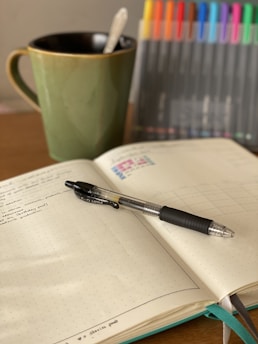 An open notebook with handwritten notes is lying on a wooden surface. A black pen rests diagonally across the notebook. To the left, a green mug with a spoon inside is positioned, and to the right, a collection of variously colored markers stands upright.