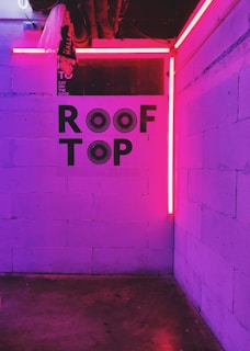 A dimly lit room with white brick walls illuminated by bright pink neon lights. The words 'ROOF TOP' are prominently displayed on the wall in bold black letters with stylized patterns. A corner display features industrial pipes and electrical wiring visible in the ceiling area.