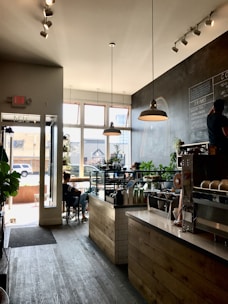 A cozy coffee shop with a rustic wooden counter and a display case filled with pastries. The shop has large windows allowing natural light to fill the space. There are several potted plants adding a touch of greenery. A chalkboard menu is on the wall listing various drinks. A few customers are seated, one near the window working on a laptop, and another at a table near the entrance.