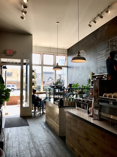 A cozy coffee shop with a rustic wooden counter and a display case filled with pastries. The shop has large windows allowing natural light to fill the space. There are several potted plants adding a touch of greenery. A chalkboard menu is on the wall listing various drinks. A few customers are seated, one near the window working on a laptop, and another at a table near the entrance.