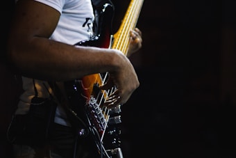 A person wearing a white shirt plays an electric bass guitar with a dark body and yellow neck. The focus is on the musician's hands and the strings of the guitar, with a dark background enhancing the prominence of the instrument.