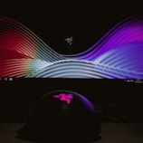 A curved display screen shows a colorful, abstract pattern with smooth wave-like lines. In front, a dark object with a pink, illuminated logo on top is visible, set against a dim background.