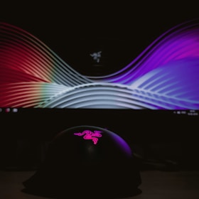 A curved display screen shows a colorful, abstract pattern with smooth wave-like lines. In front, a dark object with a pink, illuminated logo on top is visible, set against a dim background.