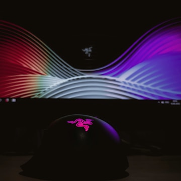 A curved display screen shows a colorful, abstract pattern with smooth wave-like lines. In front, a dark object with a pink, illuminated logo on top is visible, set against a dim background.