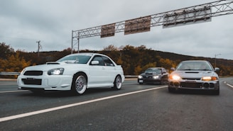 Three modified cars drive side by side on a highway. The foreground features a white car with sporty modifications and a stylized front bumper. To the right and slightly behind is a grey car with its headlights on, while in the middle and in the distance is a black car. The road is lined with autumn-colored trees against a cloudy sky.