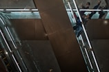 People are seen riding on a sleek, modern escalator with metal railings and glass panels. The setting appears to be indoors, possibly in a shopping mall or office building, with reflective surfaces and ambient lighting.