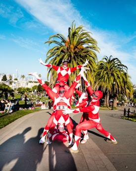 A group of individuals in red and white costumes resembling superheroes form a human pyramid in a park setting. The costumes are tight-fitting with diamond patterns, and there are tall palm trees in the background. A cityscape is visible in the distance under a clear blue sky, and people are enjoying the park surroundings.