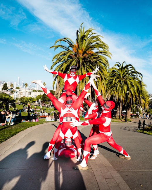 A group of individuals in red and white costumes resembling superheroes form a human pyramid in a park setting. The costumes are tight-fitting with diamond patterns, and there are tall palm trees in the background. A cityscape is visible in the distance under a clear blue sky, and people are enjoying the park surroundings.