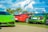 A vibrant display of luxury sports cars parked on a grassy area with palm trees in the background. The cars include a green Lamborghini, a red Porsche, and a green Audi. The sky is bright with fluffy white clouds.