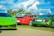 A vibrant display of luxury sports cars parked on a grassy area with palm trees in the background. The cars include a green Lamborghini, a red Porsche, and a green Audi. The sky is bright with fluffy white clouds.