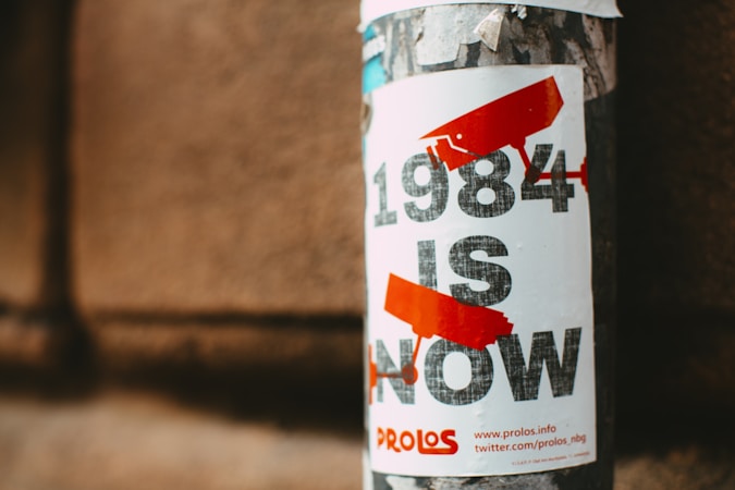 A poster on a textured wall depicts the phrase '1984 IS NOW' in bold, distressed text. Red surveillance camera icons are integrated into the design, and additional text and links are provided at the bottom of the poster.