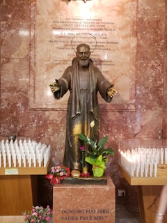 A statue of a religious figure, possibly a saint, stands in front of a marble plaque with engraved text. The statue appears to be holding its arms open in a welcoming gesture. Surrounding the statue are numerous lit candles placed on wooden stands. There are also fresh flowers and a small potted plant positioned at the base of the statue.