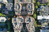 An aerial view of a residential area with multiple buildings featuring complex architectural designs. The buildings are surrounded by greenery, and the rooftops have a variety of geometric patterns. The image captures the symmetry and organization of the structures along with visible solar panels on some roofs.