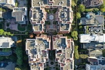 An aerial view of a residential area with multiple buildings featuring complex architectural designs. The buildings are surrounded by greenery, and the rooftops have a variety of geometric patterns. The image captures the symmetry and organization of the structures along with visible solar panels on some roofs.