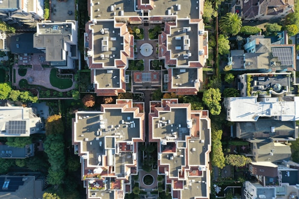 An aerial view of a residential area with multiple buildings featuring complex architectural designs. The buildings are surrounded by greenery, and the rooftops have a variety of geometric patterns. The image captures the symmetry and organization of the structures along with visible solar panels on some roofs.