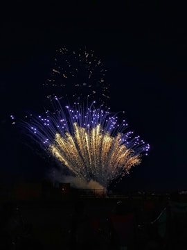 Bright and colorful fireworks burst in the night sky, with vibrant gold and blue colors lighting up the darkness. The display creates a dramatic spectacle with sparkling trails and light effects.