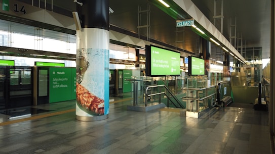 A modern train or metro station with green digital advertisements and signage in various languages. The station features clean, polished floors, glass barriers, and an escalator leading down to another level. Overhead lighting creates a bright and spacious atmosphere.