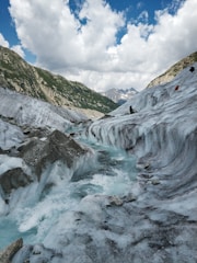 An icy glacier flows through rugged mountain terrain under a partly cloudy sky. The ice is a mix of white and light blue, with visible crevasses and ridges. There are mountains covered with greenery in the background and patches of snow.