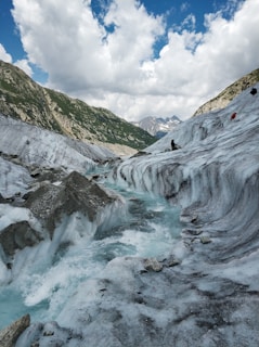 An icy glacier flows through rugged mountain terrain under a partly cloudy sky. The ice is a mix of white and light blue, with visible crevasses and ridges. There are mountains covered with greenery in the background and patches of snow.