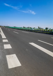 A long, open highway with white lane markings stretches under a clear blue sky. Green railings line the side of the road, with trees and small shrubs visible beyond them on a flat landscape.