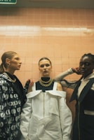 Three fashionable individuals stand in front of a tiled wall, each wearing unique and stylish clothing. The central figure wears a striking white outfit with a large gold chain necklace. The person on the left sports a patterned jacket and gold hoop earrings, while the individual on the right is dressed in dark clothing with a gold chain. The lighting is subdued, enhancing the dramatic effect.