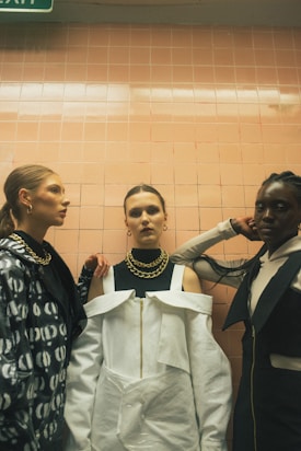 Three fashionable individuals stand in front of a tiled wall, each wearing unique and stylish clothing. The central figure wears a striking white outfit with a large gold chain necklace. The person on the left sports a patterned jacket and gold hoop earrings, while the individual on the right is dressed in dark clothing with a gold chain. The lighting is subdued, enhancing the dramatic effect.