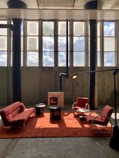 A spacious industrial-style room features tall windows with sunlight streaming in, casting shadows on the concrete floor. In the center, a setup of modern red furniture includes a sofa, two armchairs, and a plush red rug. Two small black tables and a contemporary standing lamp complement the minimalist aesthetic.