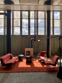 A spacious industrial-style room features tall windows with sunlight streaming in, casting shadows on the concrete floor. In the center, a setup of modern red furniture includes a sofa, two armchairs, and a plush red rug. Two small black tables and a contemporary standing lamp complement the minimalist aesthetic.