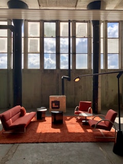 A spacious industrial-style room features tall windows with sunlight streaming in, casting shadows on the concrete floor. In the center, a setup of modern red furniture includes a sofa, two armchairs, and a plush red rug. Two small black tables and a contemporary standing lamp complement the minimalist aesthetic.