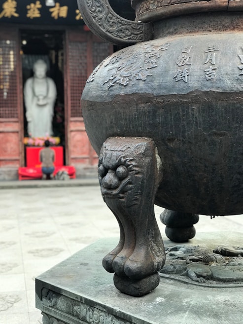 An ornate, large bronze vessel with intricate engravings and design stands prominently in the foreground. The leg of the vessel features a sculpted lion or mythological creature. In the background, a person kneels in front of a white statue inside a temple, shrouded in a spiritual ambiance, with a red altar adorned with various offerings.