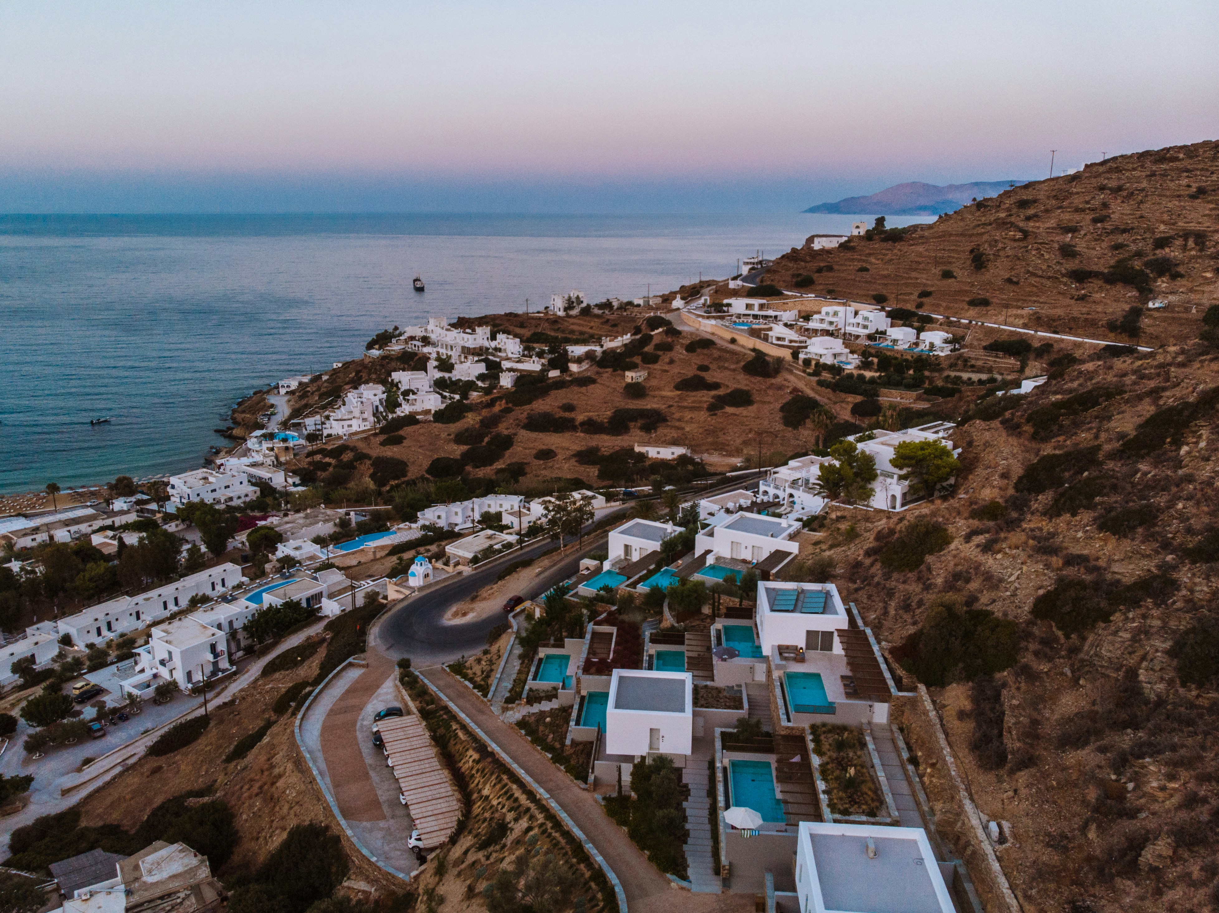 Elevated luxury hillside villa development with panoramic Aegean sea views, modern white architecture, large glazing and infinity pool at sunset