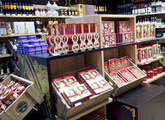 Shelves filled with a variety of wine bottles and boxes of chocolates, prominently featuring red and gold packaging. An array of colorful liquor bottles and gift boxes are also displayed, emphasizing a luxurious and premium shopping atmosphere. The architectural design on the shelving adds a sophisticated touch.