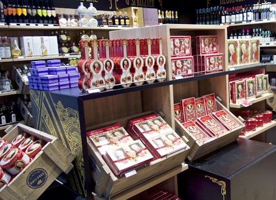 Shelves filled with a variety of wine bottles and boxes of chocolates, prominently featuring red and gold packaging. An array of colorful liquor bottles and gift boxes are also displayed, emphasizing a luxurious and premium shopping atmosphere. The architectural design on the shelving adds a sophisticated touch.