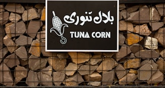 A sign with the words 'TUNA CORN' written in English and another language on a black background. The sign features an image of corn and is mounted on a stone wall held together by a metal frame.