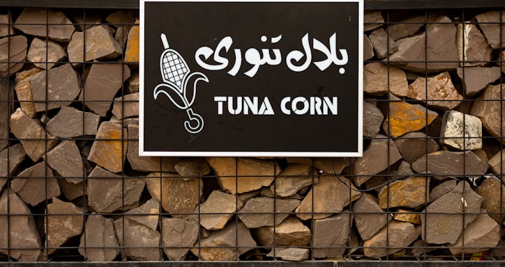 A sign with the words 'TUNA CORN' written in English and another language on a black background. The sign features an image of corn and is mounted on a stone wall held together by a metal frame.