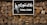 A sign with the words 'TUNA CORN' written in English and another language on a black background. The sign features an image of corn and is mounted on a stone wall held together by a metal frame.