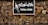 A sign with the words 'TUNA CORN' written in English and another language on a black background. The sign features an image of corn and is mounted on a stone wall held together by a metal frame.