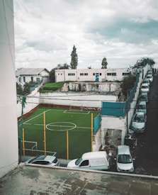 A small sports field with green artificial turf is enclosed by a high net fence. Vehicles, including a white van and several cars, are parked around the perimeter. A set of buildings, one of which appears dilapidated, is visible in the background. The sky is overcast, suggesting gloomy weather.