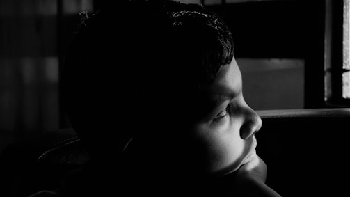A black and white portrait of a child gazing thoughtfully out a window, soft natural light framing their face.