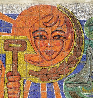 A mosaic art piece featuring a stylized face with a laurel wreath surrounded by vibrant colors. The face appears to be smiling and is holding a key, depicted with intricate tile work. The background includes shades of blue and green, adding to the dynamic composition.