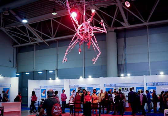 A red robotic structure resembling a large mechanical spider is suspended from the ceiling in a convention or exhibition hall. Below, a group of people is gathered, some observing the robot and others walking around. The walls display booth numbers and names, indicating trade show or conference stalls.