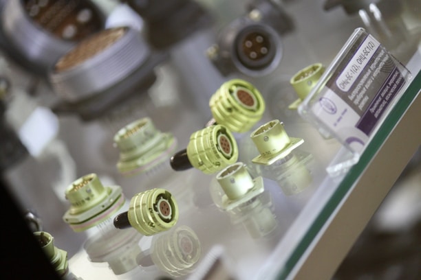 A collection of various electrical connectors is displayed in a glass case. These connectors come in multiple sizes and shapes, primarily in a light green color with metal pins. Some connectors have a visible black section, possibly a protective cap or housing. An informational card is positioned next to the connectors on the right side.