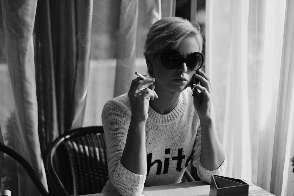 grayscale photography of woman using phone while holding cigarette sitting near table