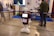 A humanoid robot with green-lit eyes and a screen on its chest displaying a logo stands in an exhibition space. It is surrounded by various pieces of equipment and people interacting with different technology displays. The setting appears to be industrial with smooth flooring, tables, chairs, and informational posters on the wall.