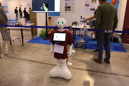 A humanoid robot with green-lit eyes and a screen on its chest displaying a logo stands in an exhibition space. It is surrounded by various pieces of equipment and people interacting with different technology displays. The setting appears to be industrial with smooth flooring, tables, chairs, and informational posters on the wall.