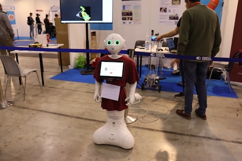 A humanoid robot with green-lit eyes and a screen on its chest displaying a logo stands in an exhibition space. It is surrounded by various pieces of equipment and people interacting with different technology displays. The setting appears to be industrial with smooth flooring, tables, chairs, and informational posters on the wall.
